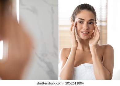 Daily Skin Care. Millennial Lady Touching Face With Smooth Skin Looking At Her Reflection In Mirror Standing In Bathroom After Shower. Pampering And Skin Care Concept. Selective Focus