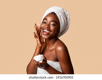 Daily Skincare Concept. Portrait of beautiful young african american woman holding cream jar and applying moisturizer on cheek, looking at camera. Smiling black lady wearing white towel on head