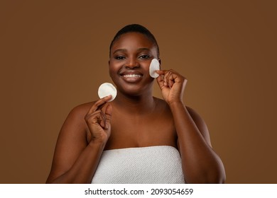 Daily Skincare Routine. Chubby Black Woman Wrapped In White Towel Cleaning Face With Cotton Pads, Using Toner Or Lotion, Standing Posing Isolated On Brown Studio Background, Looking At Camera