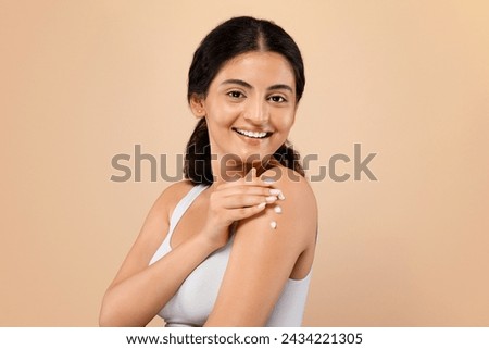 Daily Skincare. Young indian woman with pleasant smile gently applying dab of cream on her shoulder, attractive eastern lady illustrating beauty routine, standing against beige studio background