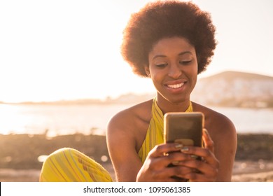 Daily Summer Sunnny Scene With Black Race Afro American Beautiful Girl With Alternative Hair Looking At The Phone And Check Social Media. Sunny Golden Light Beach And Ocean In The Background. 