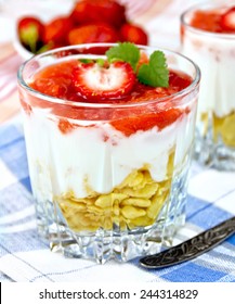 Dairy dessert with strawberries, corn flakes and yogurt in two glassful, spoon, blue napkin, strawberriy in a bowl on linen tablecloth background