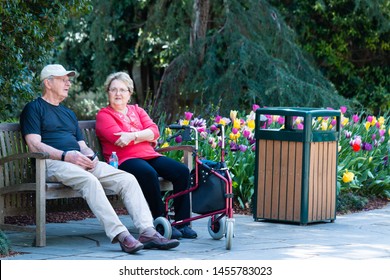 Dallas, Texas/USA - May 2019: Senior Citizens - Man and Woman with Walker Sitting on Bench Resting and Talking Flowers in Background Trash Can on side