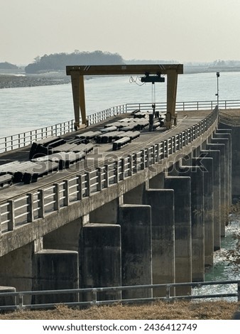 A dam gate with a heavy-duty gantry crane overlooking a river. The structure has concrete pillars supporting the barrier, with metal counterweights positioned on the gate.