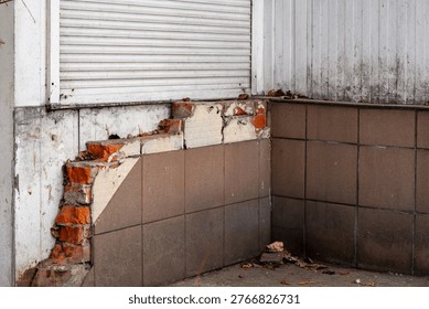 Damaged building facade with falling ceramic tiles and exposed red bricks, destroyed exterior wall needing renovation, old structure with white roller shutter, architectural decay and construction mat