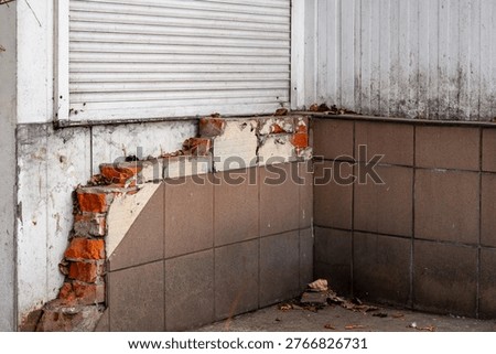 Damaged building facade with falling ceramic tiles and exposed red bricks, destroyed exterior wall needing renovation, old structure with white roller shutter, architectural decay and construction mat