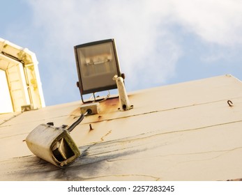 Damaged CCTV camera hanging on a cable on yellow wall, old flood light above the camera. Front element is broken and there is sign of explosion. Blue cloudy sky. War or high criminal unsafe area. - Powered by Shutterstock