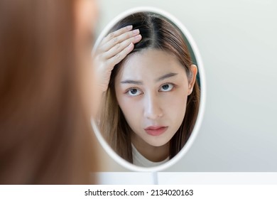 Damaged Hair, face serious asian young woman worry looking at scalp in mirror, hand in break into front hair loss, thin problem symptom at home. Health care shampoo beauty, isolated on background. - Shutterstock ID 2134020163