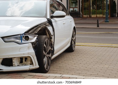 Damaged Left Front Side Of A White Car Parked On The Street: No Fender, Shattered Headlight, Broken Bumper, Damaged Frame, No Fog Light. 
