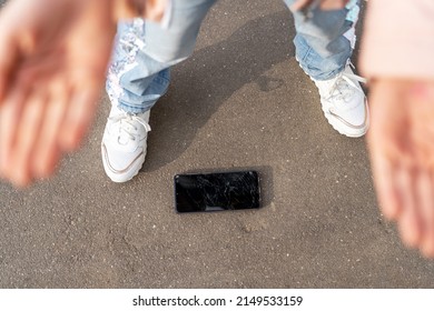 Damaged mobile phone with cracked touch screen. Elevated view of foot and hands with broken screen smartphone dropped on floor on asphalt concept. 