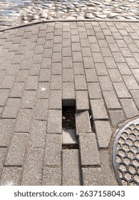 Damaged sidewalk paving with missing bricks near manhole cover. Urban infrastructure issue creating a trip hazard. Daylight photo of cracked pavement surface in public space.