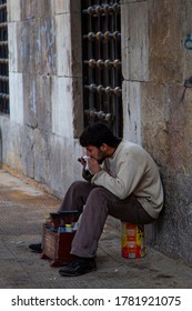 Damascus, Syria 03/28/2010: A Poor Young Syrian Man In Torn Clothes Is Sitting On An Empty Can  In Front Of His Shoe Shine Stand. He Is Taking A Bite From His Lunch With His Shoe Dye Stained Hands.