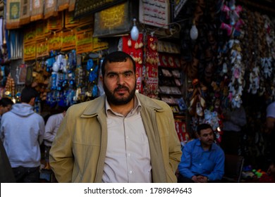 Damascus, Syria 03/28/2010: Selective Focus Portrait Of A Bearded Syrian Shopkeeper In Al Hamidiyah Souq As He Stands In Front Of  Displays Of Shops Selling Local Ornaments, Clothings And Souvenirs.