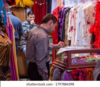 Damascus, Syria 03/28/2010: A Shop Keeper In Al Hamidiyah Souq Is Reading Newspaper While Standing At The Entrance Of His Boutique. He Has Traditional Colorful Long Woman Dresses And  On Display.