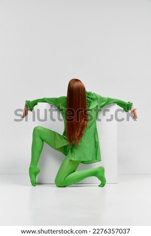 Dance. Avant-garde and experimental art style of human portrait. Unknown woman with long glossy red hair in green outfit posing over light background. Body language, faceless emotions concept