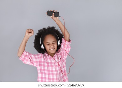 Dance! Little girl in headphones holding mobile phone and dancing with closed eyes on grey background