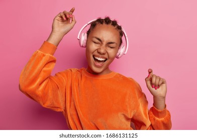 Dance With Me. Upbeat Dark Skinned Teenager Raises Arms, Moves With Rthythm Of Melody, Feels Energetic, Dressed In Bright Orange Jumper, Has Fun, Isolated On Pink Background. Music Brings Joy