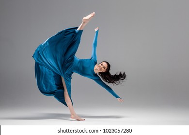 Dance split position of beautiful happy brunette ballerina in blue evening dress smiling at camera, motion studio shot, copy-space. Grey background