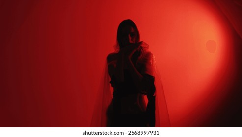 Dance therapy. Calming practice. Peaceful woman silhouette in cellophane rags performing hypnotic hand movements isolated on dark red glowing copy space background.