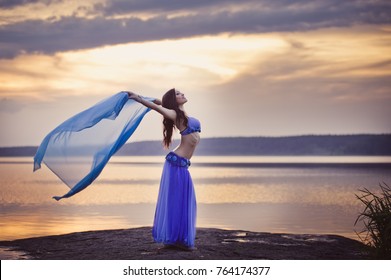 Dancer of bellydance in a blue suit on the beach, against the background of the water. Beautiful nature at sunset. Belly dance. Girl with exotic appearance. Oriental beauty.