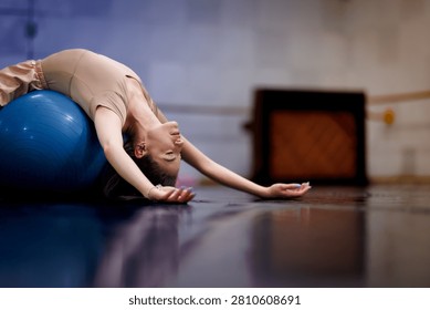 A dancer practices by lying back on a blue exercise ball. The setting is a dance studio with wooden floors. The space is designed for dance training and rehearsal.