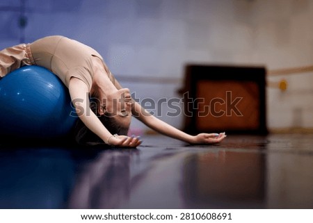 A dancer practices by lying back on a blue exercise ball. The setting is a dance studio with wooden floors. The space is designed for dance training and rehearsal.