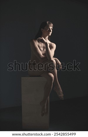 The dancer sits thoughtfully on a stool, her body illuminated by soft studio light. The nude tones of her attire and the delicate shadows create a serene and introspective mood
