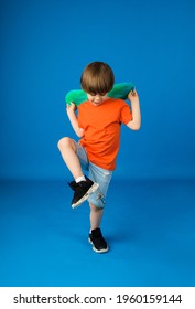 Dancing Boy With Brown Hair In An Orange T-shirt And Denim Shorts Holds A Skateboard On A Blue Background With Space For Text