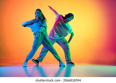 Dancing Couple. Stylish Man And Woman Dancing Hip-hop In Casual Youth Clothes On Gradient Yellow Red Background At Dance Hall In Neon Light. Youth Culture, Hip-hop, Fashion, Breakdance, Action And Ad