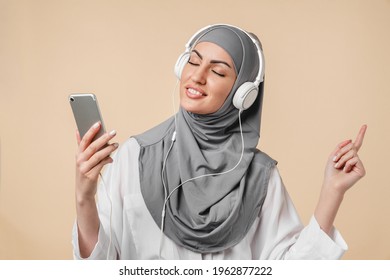 Dancing happy young muslim middle eastern islamic arabian woman student listening to the music on smart phone and headphones headset over beige background. Cheerful woman in grey hijab dancing - Powered by Shutterstock