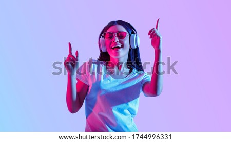 Dancing in modern wireless headphones. Asian girl with glasses enjoys favorite song, isolated in neon, studio shot