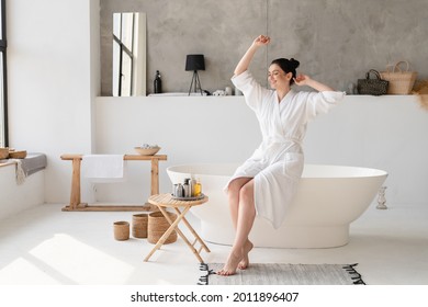 Dancing Young Caucasian Woman In White Spa Bathrobe Relaxing Stretching In Bathroom Before Taking Shower Bath At Home. Beauty Treatment