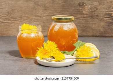 Dandelion jam or honey in the glass jar with fresh blooming dandelion flowers. Seasonal product, trendy hard light, dark shadow. Stone concrete background, close up