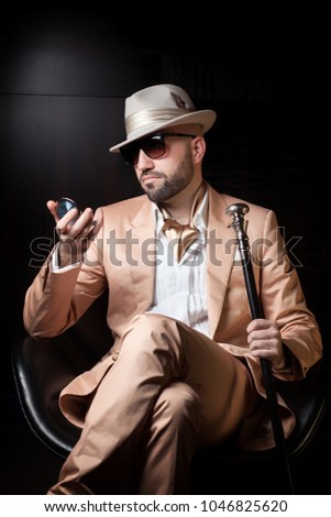 Dandy man dressed as mafia boss, pimp or playboy looks at himself in the pocket mirror, wearing sunglasses, white hat and pink or peach suit, sitting in a leather chair, isolated on black background 