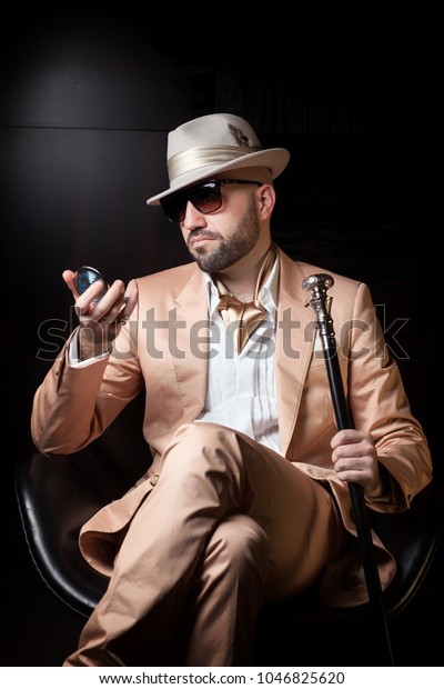 マフィアのボス ポンプ プレイボーイの格好をしたダンディな男性が 黒い背景に革の椅子に座り サングラス 白い帽子 ピンクまたは桃のスーツを身に着けて ポケットミラーの中を見る の写真素材 今すぐ編集