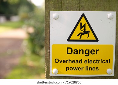 Danger Overhead Electrical Power Lines Warning Sign On Wooden Post With Out Of Focus Background