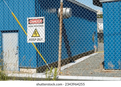 A danger sign, high voltage, with keep out electricity symbol hanging on a mesh wire fence with two blue building in an enclosed yard. The electrical station storage building has a single grey door. 