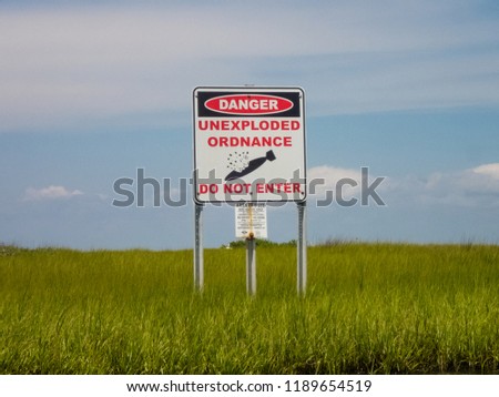 Danger unexploded ordnance do not enter  posted sign above green sea grass in front of a blue sky wide view