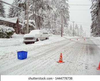 Dangerous Downed Power Lines During A Winter Storm. Power Lines Are On Top Of A Snowed In Car And On The Snow Covered Street. A Car Is Approaching. Traffic Cones Are Placed Around The Area.