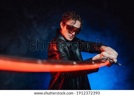 Dangerous and martial guy dressed in black leather clothing with sunglasses poses in blue background holding sword.
