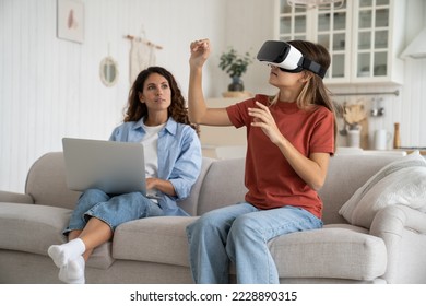 Dangers of virtual reality for children. Curious schoolgirl daughter using virtual reality simulator headset for study or entertainment while mother working remotely on laptop. Technology and family - Powered by Shutterstock