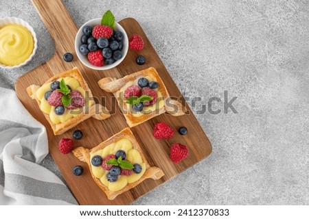 Danish puff pastry cakes with custard cream and fresh berries, sprinkled with powdered sugar on a wooden board on a concrete background. Top view, copy space