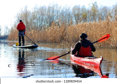 DANUBE RIVER, UKRAINE - DECEMBER 09, 2017: Man Paddles A Red Kayak And Man Standing With A Paddle On The Stand Up Paddle Board (paddleboard, SUP) On The River Or Lake In Fall Season. Winter Kayaking