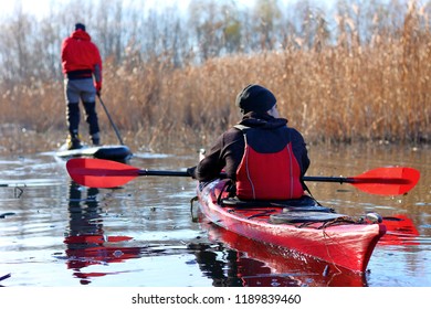 DANUBE RIVER, UKRAINE - DECEMBER 09, 2017: Man Paddles A Red Kayak And Man Standing With A Paddle On The Stand Up Paddle Board (paddleboard, SUP) On The River Or Lake In Fall Season. Winter Kayaking