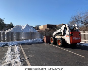 Danvers, MA / USA - Jan 2020: Compact Bobcat Tractor After Cleaning A Parking Lot With A Huge Pile Of Snow In The Background