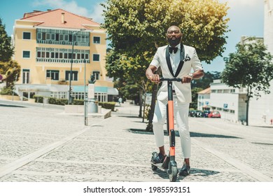 A dapper adult bearded bald black guy in eyeglasses and a white fashionable summer costume is holding in hands the handlebars of the e-scooter ready to ride over the paving-stone on a warm sunny day