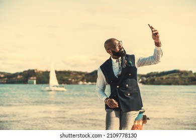 A dapper African-American senior in a fashionable outfit is sitting on a bollard next to the river and having video call on a dazzling day; an elegant black guy in a costume is taking a selfie on pier