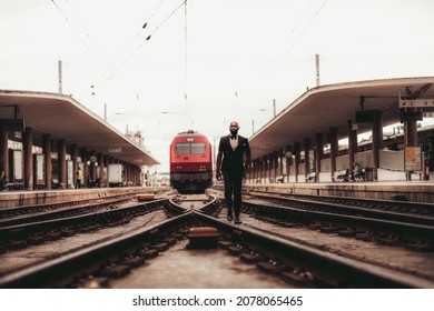 A dapper bald bearded African man in a fashionable suit with a bow-tie is going towards the camera along the symmetrical railway switch with a red locomotive train in between two railroad platforms