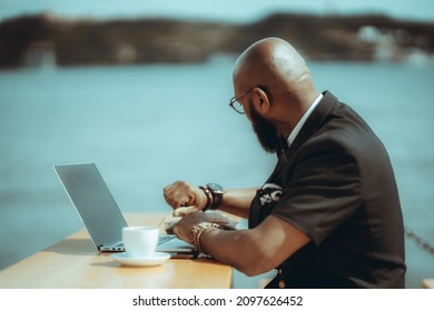 A dapper bald, bearded black guy with glasses looks at his watch. A handsome businessman is waiting for a person late for a meeting at a coffee shop. An elegant man alone at an outdoor table