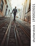 A dapper bald bearded guy in an elegant custom-made business costume and eyeglasses is leaning on his walking stick while staying in the middle of a narrow antique European street on the tramway track